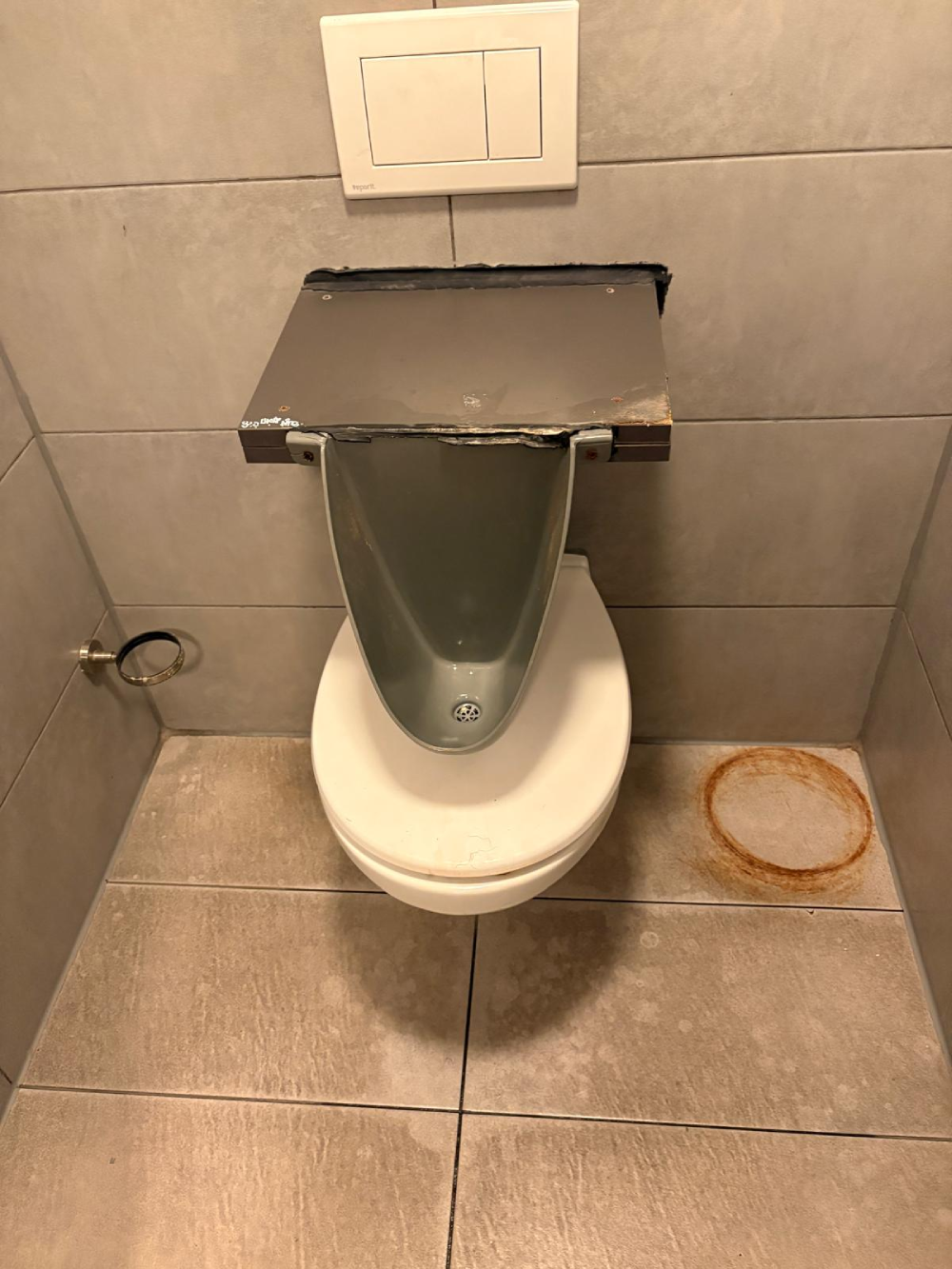 A urinal installed directly above a white toilet bowl, with a circular rust stain on the floor nearby.