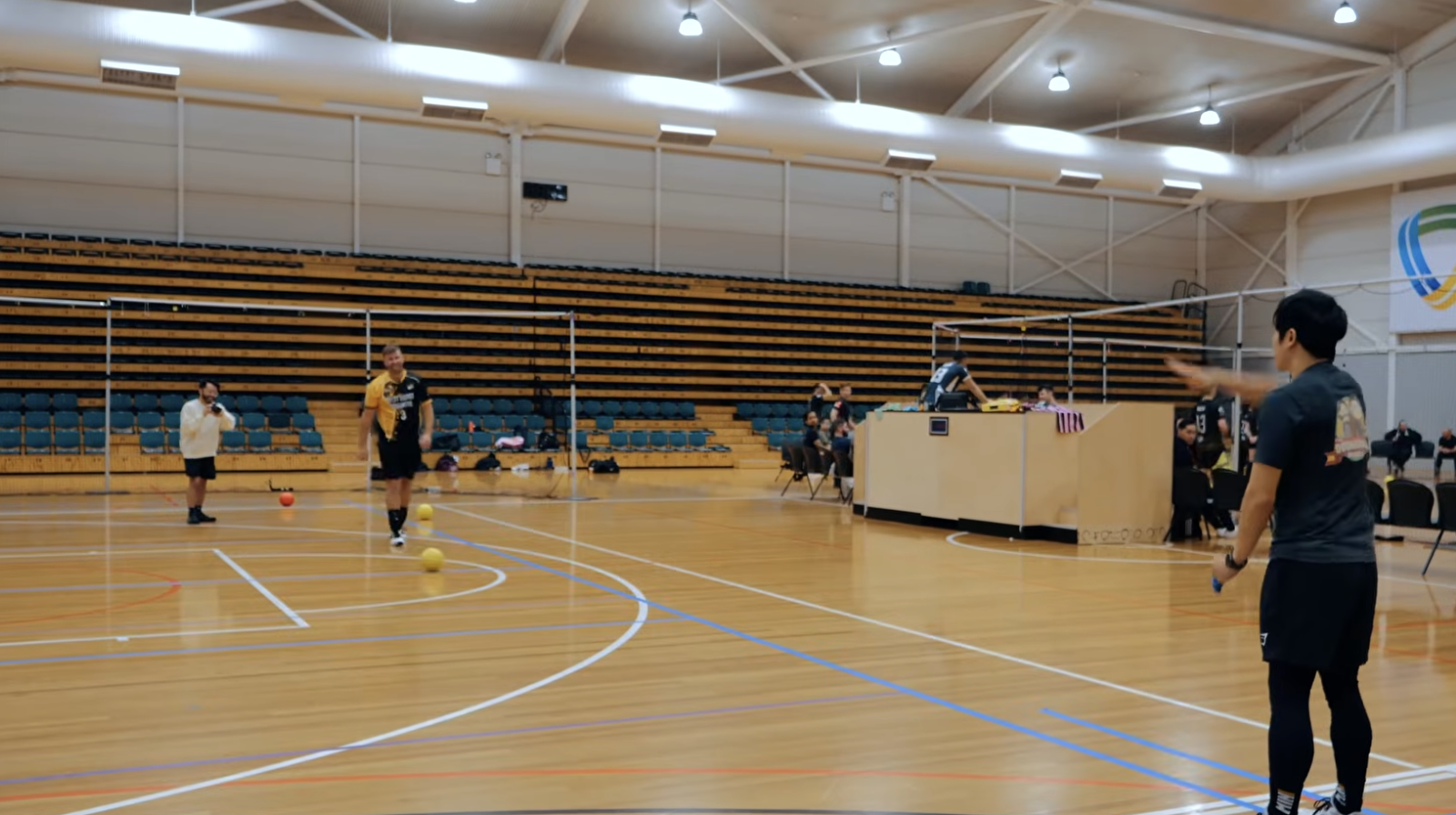 A volleyball court with a few players standing on the floor in front of rows of empty stadium seating.