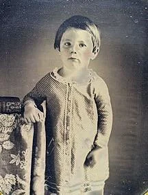 A portrait of a young child in a long-sleeved tunic standing beside a patterned chair.