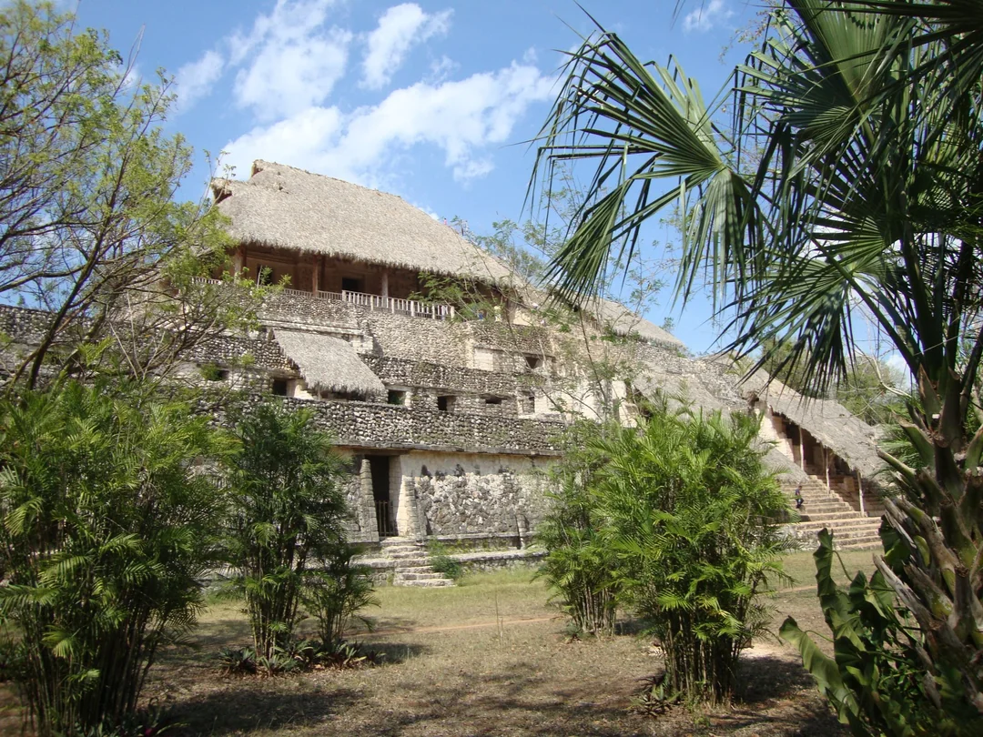 Large stone Mayan pyramid with a thatched roof, surrounded by trees and palm plants.