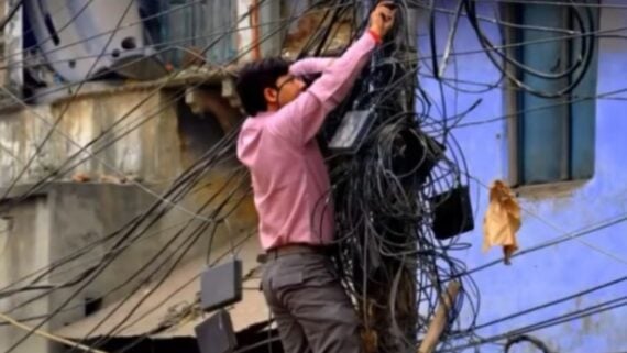 A man in a pink button-down shirt stands on a pole to work on a large, tangled cluster of utility wires.