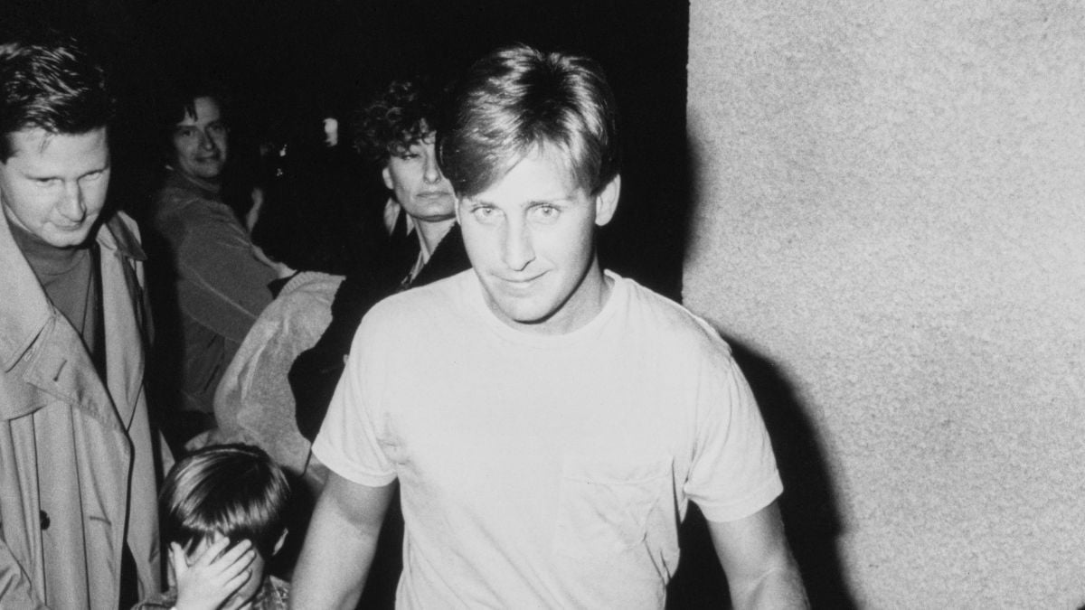 A man in a white t-shirt walks toward the camera in a black-and-white photo, with several people behind him, including a child covering their face.
