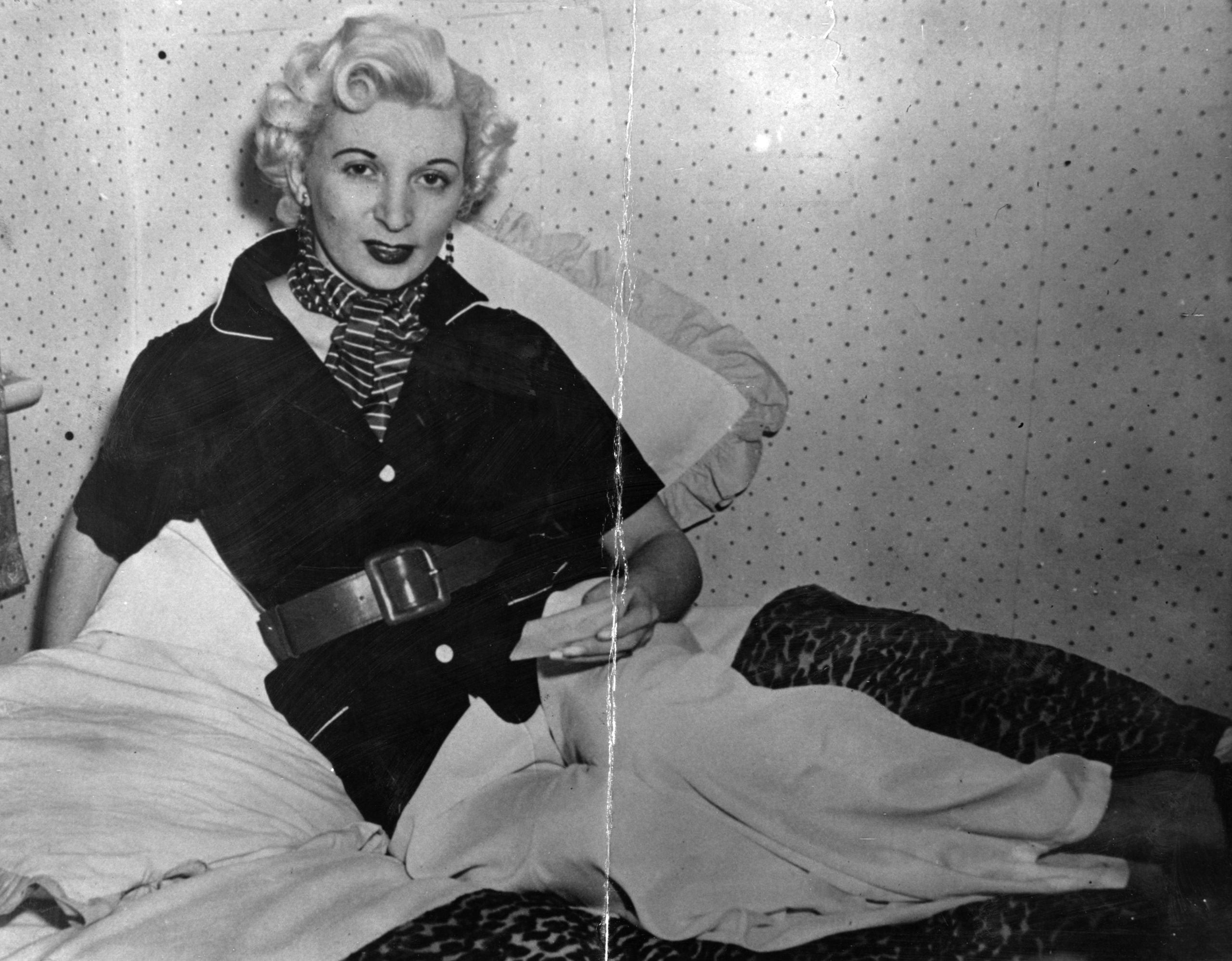 A blonde woman with a striped scarf and belted dark shirt reclining on a bed.