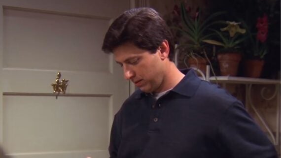 A man with short dark hair wearing a black polo shirt looks down thoughtfully. He stands in front of a beige door with a star-shaped ornament and some potted plants in the background.