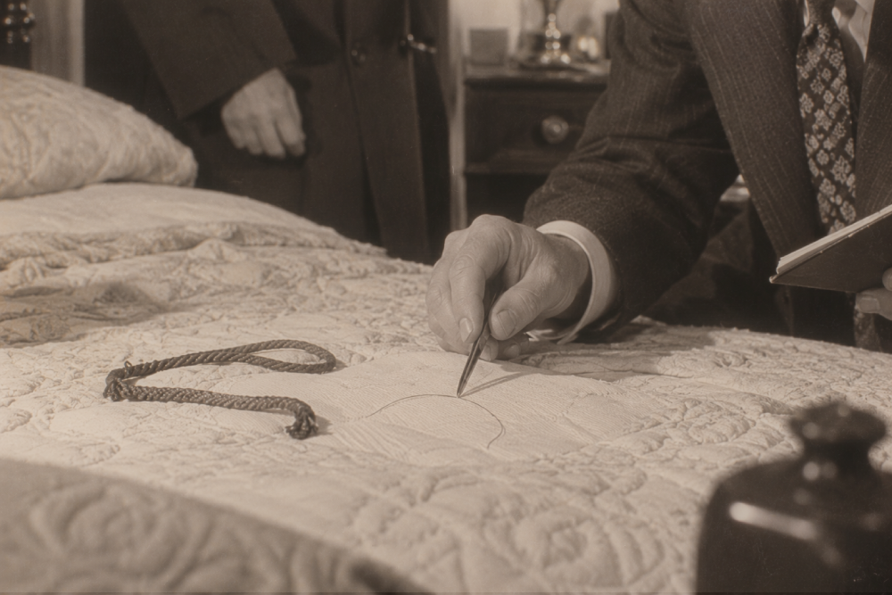 A person in a suit uses a pen to trace a circular design onto a quilted bedspread near a piece of cord.