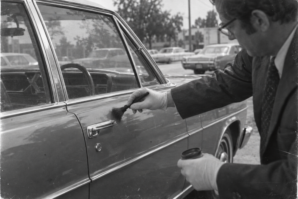 A person in a suit and gloves uses a brush to dust a car door handle for fingerprints.