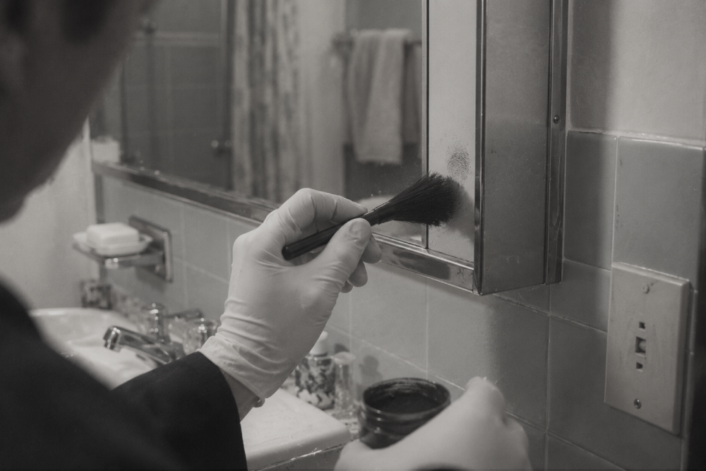 A person wearing a glove brushes dusting powder onto a fingerprint on a bathroom medicine cabinet mirror.
