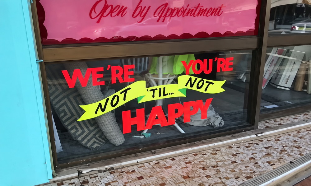 Store window sign reading “Open by Appointment” above red and yellow lettering that says “We’re not happy ‘til you’re not happy.”