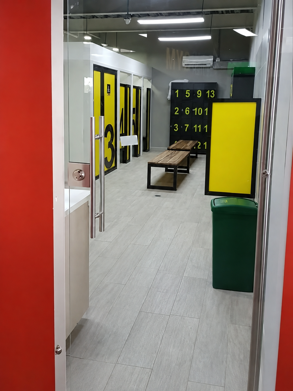 View through a glass doorway into a locker room featuring rows of yellow-numbered lockers, wooden benches, and a green trash bin.