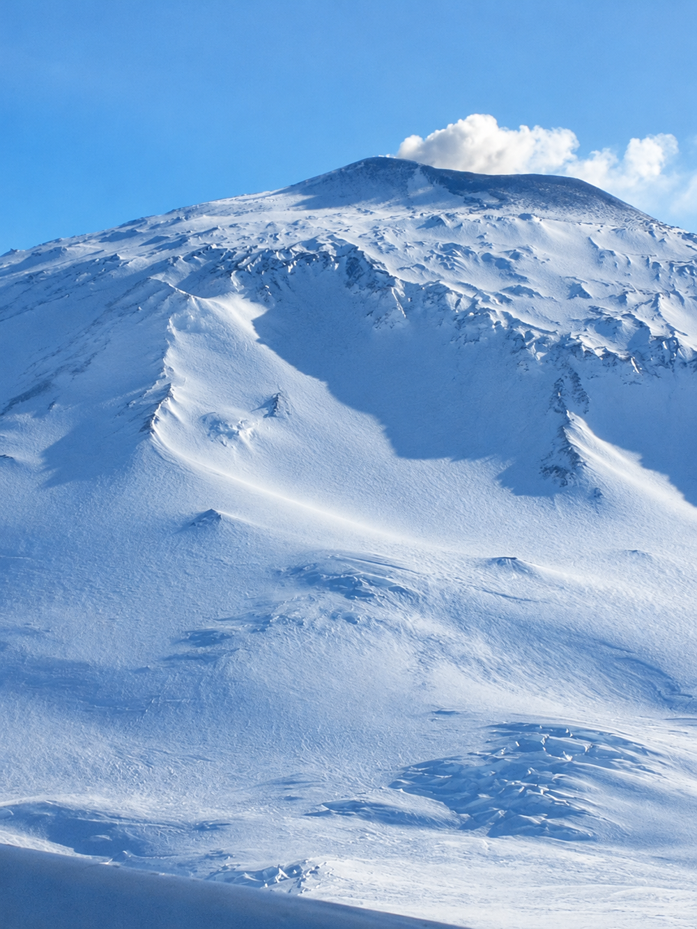 A snow-covered volcanic mountain peak with a plume of smoke rising from the summit.