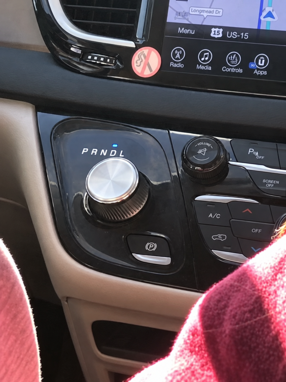 Car interior console showing a rotary gear shifter with PRNDL indicators, a volume knob, and a parking brake button below a touchscreen display.