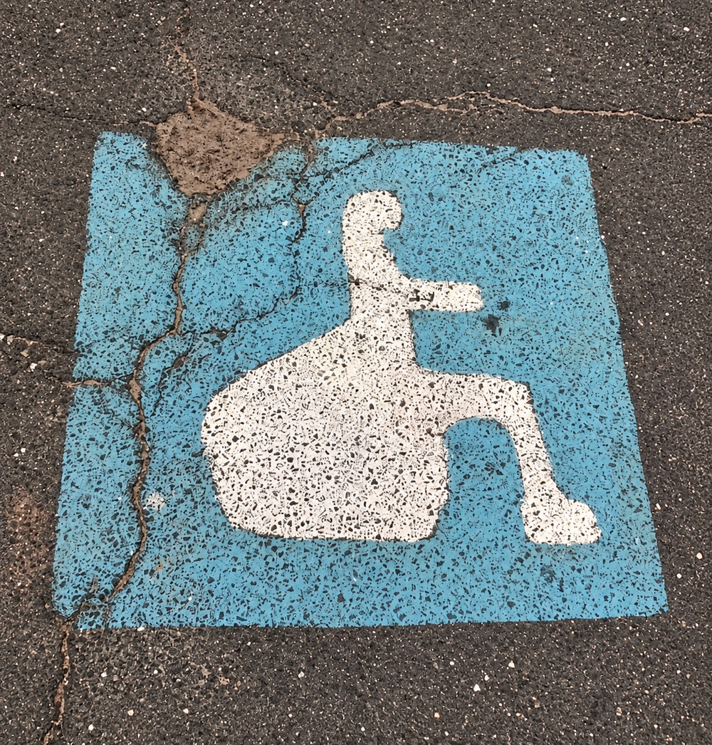 A white accessibility symbol painted on a cracked blue square on asphalt.