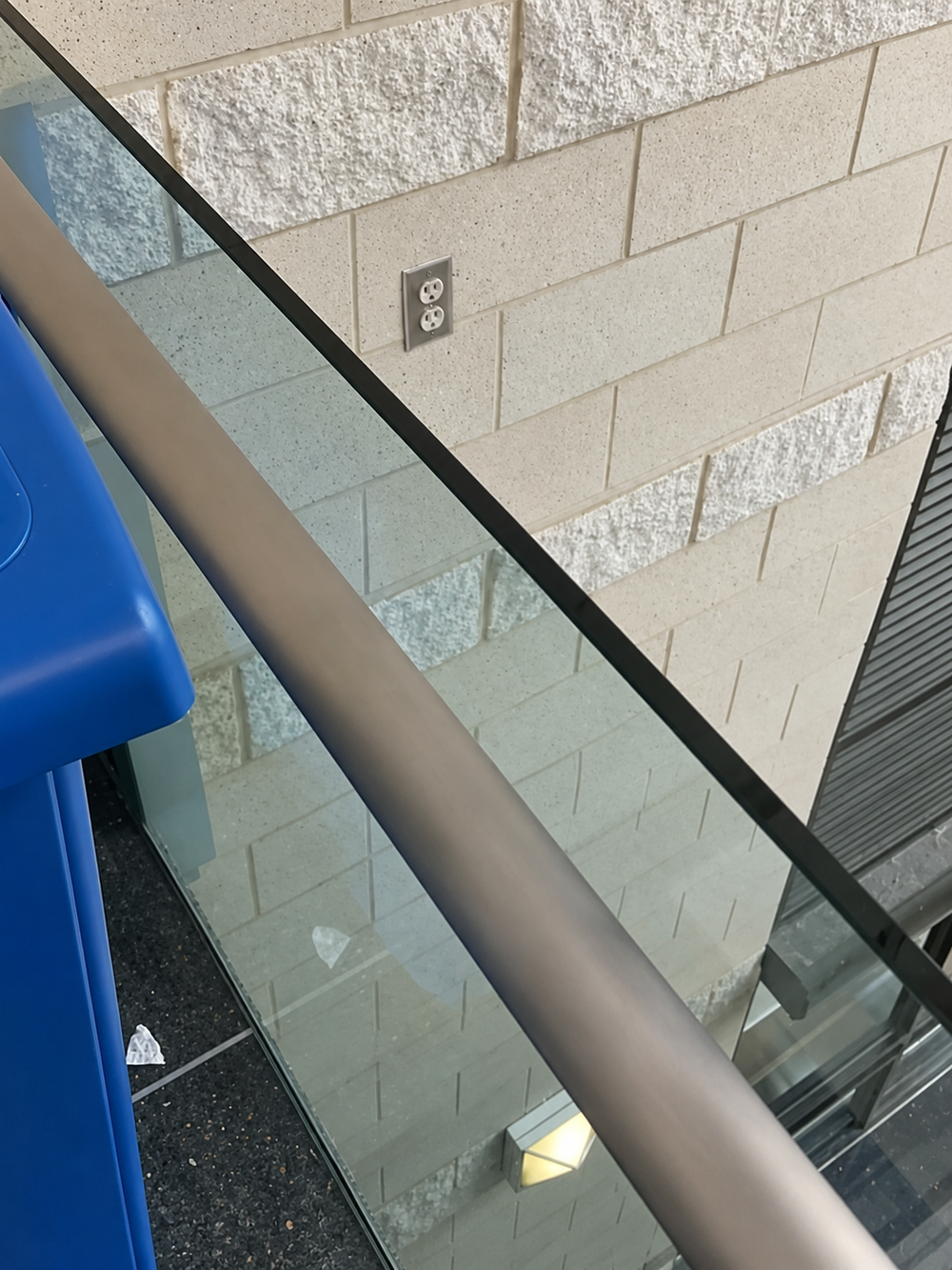 A glass railing with a metal handrail in front of a white brick wall featuring a wall outlet.