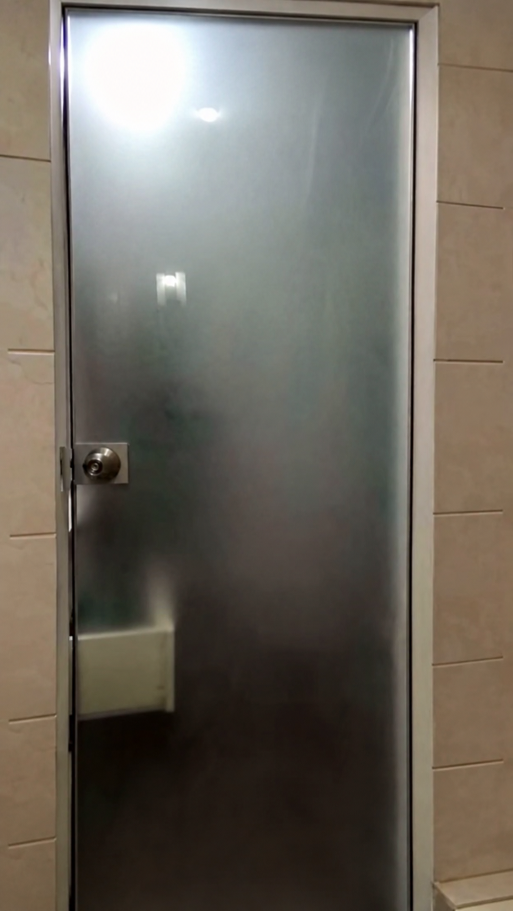 A frosted glass door with a silver frame and a round door knob, set into a tiled wall.