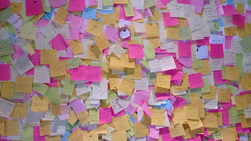 A wall covered in many overlapping square sticky notes in shades of pink, yellow, blue, and white, with some notes featuring handwritten text and a small bear-shaped cutout.