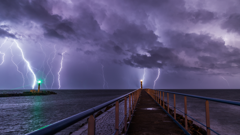 A stone pier leads toward a lighthouse under a dark, stormy sky filled with lightning strikes.