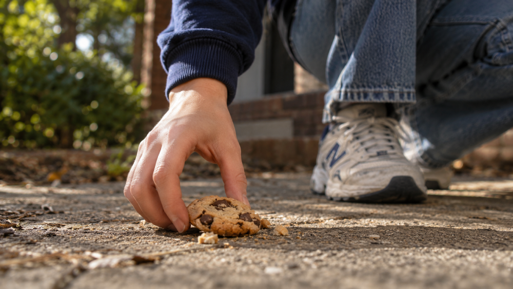 A person crouching on a sidewalk reaches down to pick up a chocolate chip cookie from the ground.