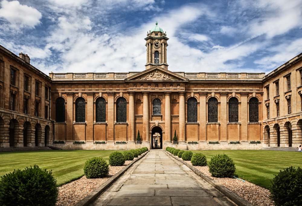 A stone courtyard with a central path leading to a classical building featuring a clock tower under a cloudy sky.