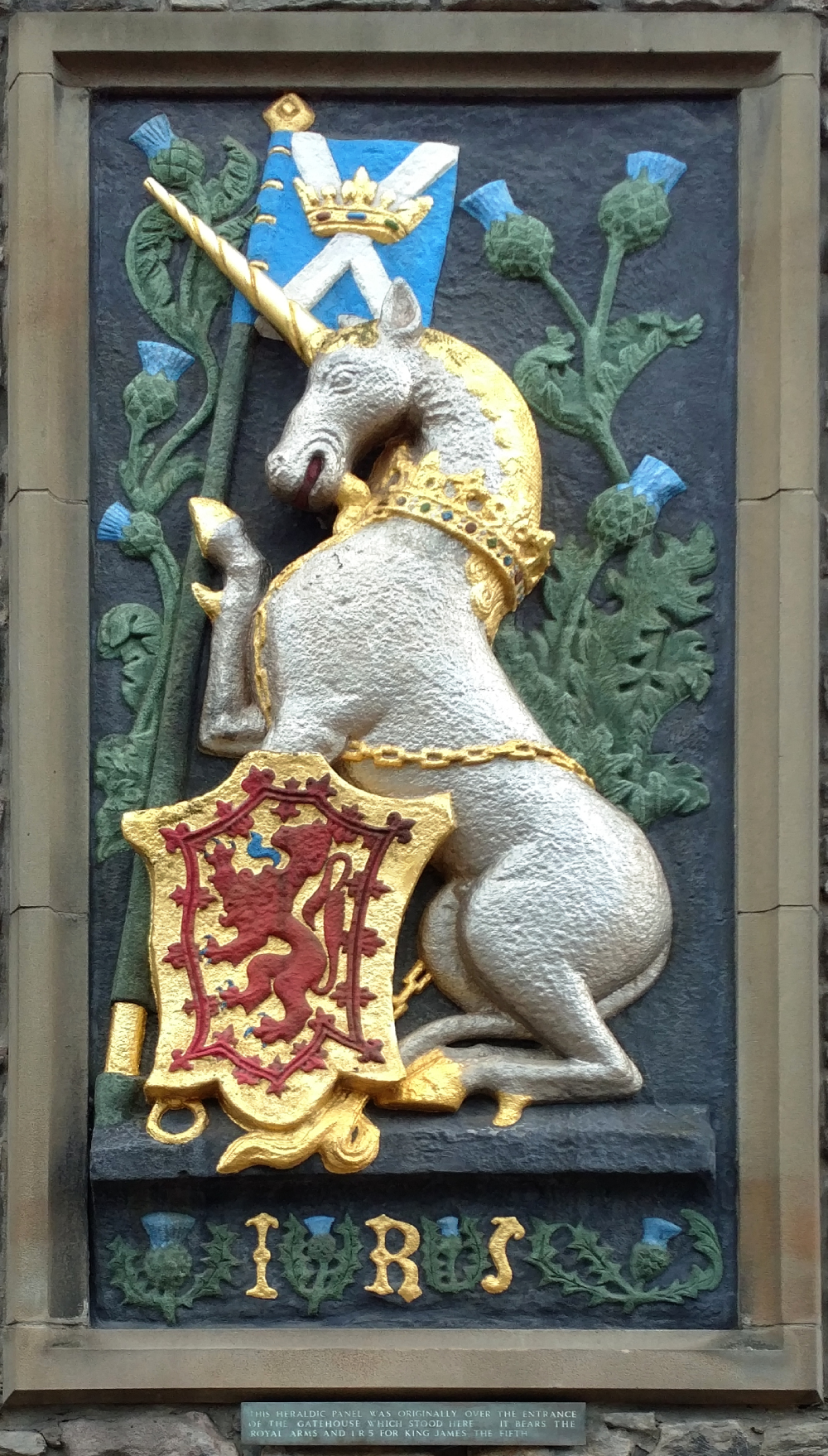 A stone relief carving of a crowned, silver unicorn holding a banner with a blue and white saltire, standing beside a shield featuring a red lion rampant, with the letters I R 5 below and thistle plants flanking the scene.