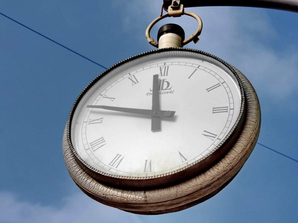 A large, round, outdoor analog clock with Roman numerals hangs against a blue sky.