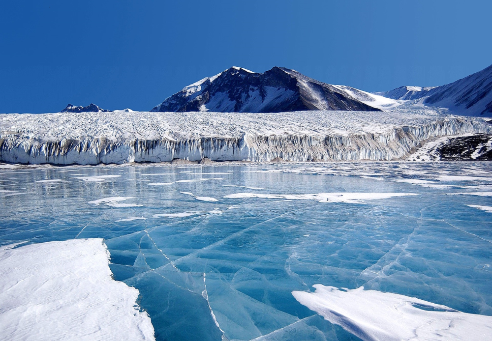 A vast glacier flows into a lake covered by thick, cracked blue ice with snowy peaks in the background.