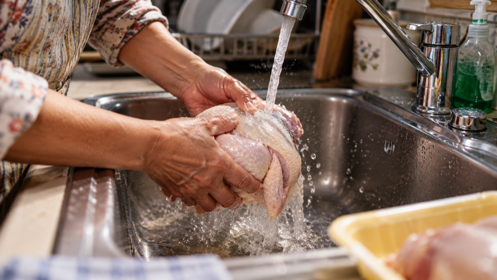 A person rinsing a raw whole chicken under a kitchen faucet.