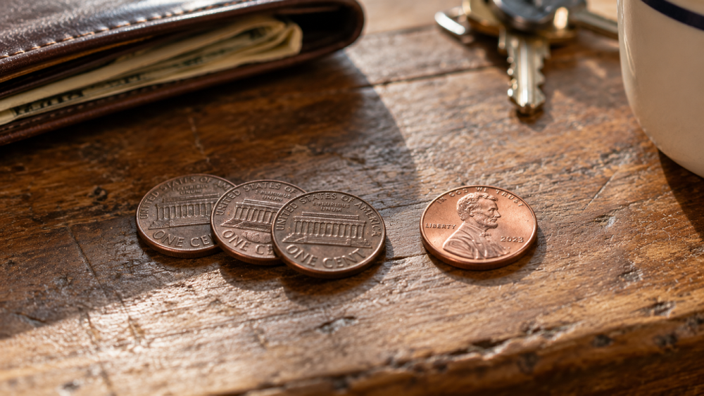 Four US pennies, a leather wallet containing paper money, and a set of keys on a wooden surface.
