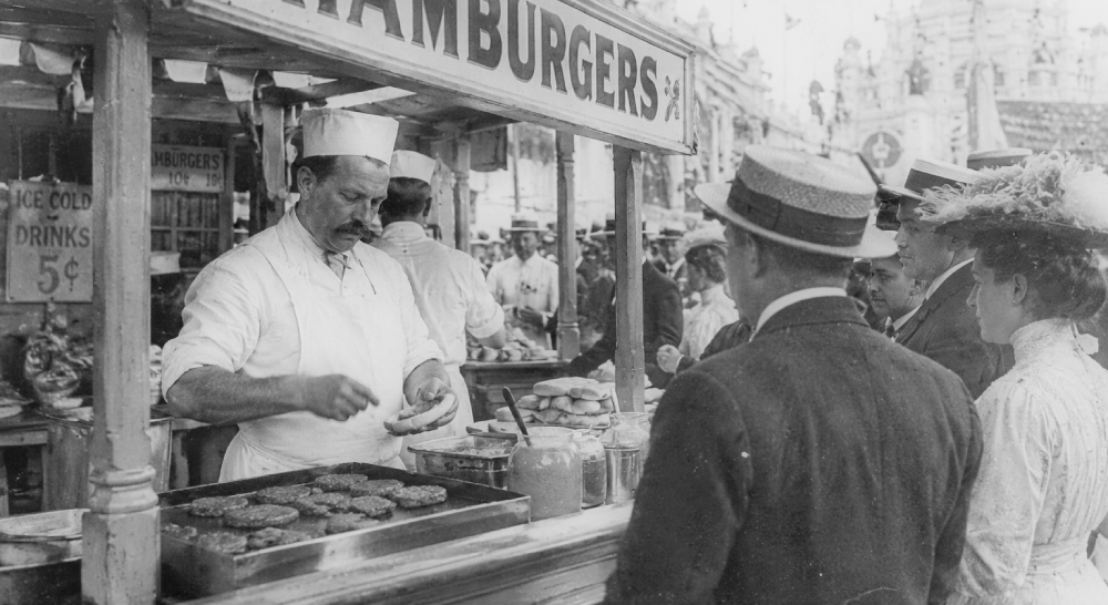 A man in a white apron and cap cooks hamburgers at a street stall for a crowd of customers wearing period hats and clothing.