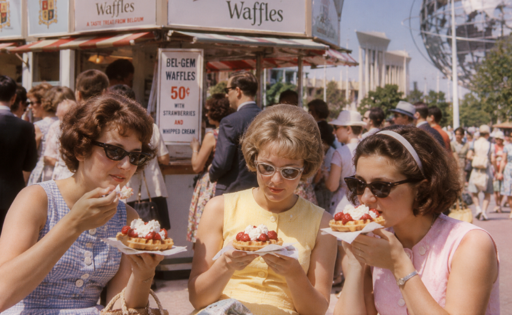 Three women in vintage clothing eat waffles topped with strawberries and whipped cream in front of a fairground booth.