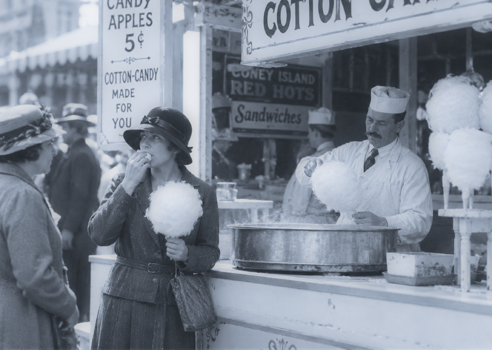 A woman eating cotton candy stands at a carnival booth while a vendor prepares more spun sugar nearby.
