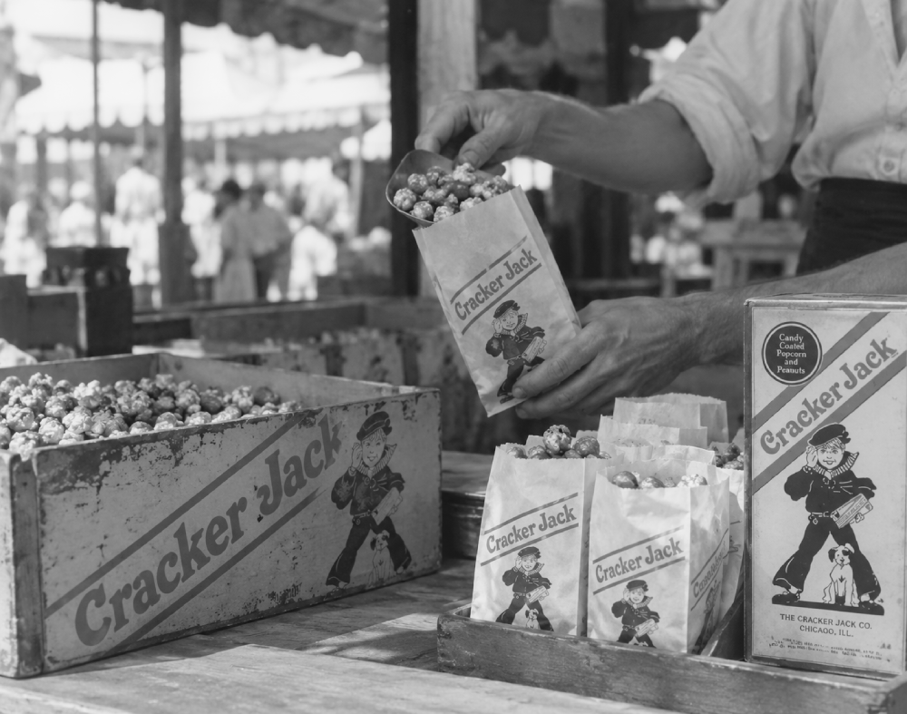 A person filling paper bags with Cracker Jack popcorn from a wooden crate at a stand.
