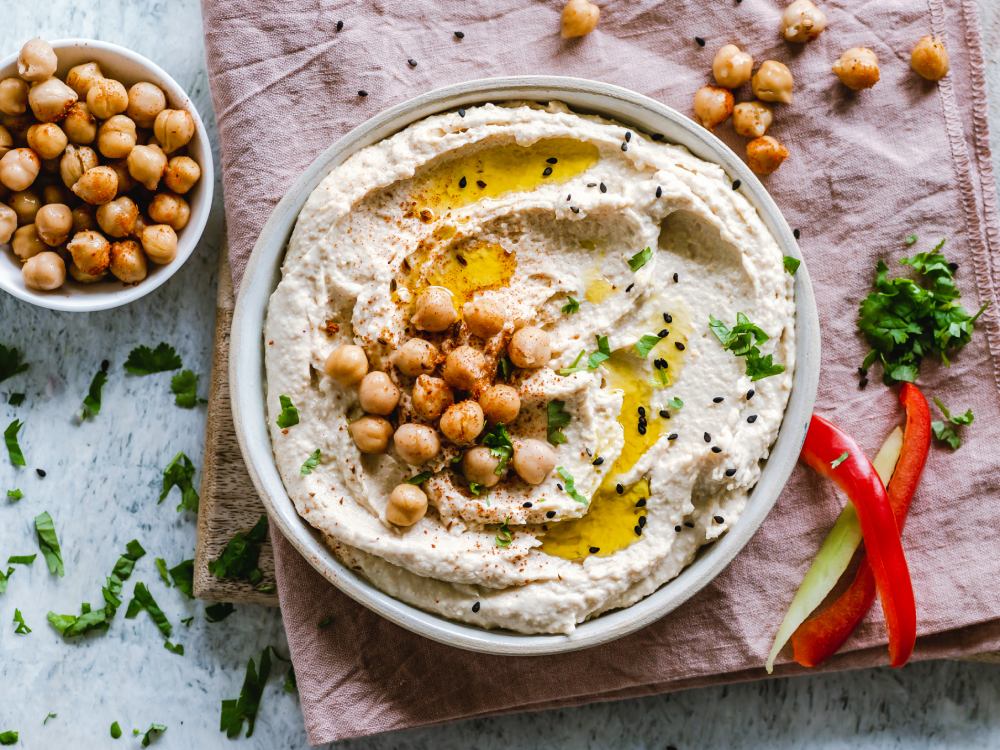 A bowl of hummus topped with chickpeas, olive oil, and spices, served with a side bowl of chickpeas, red bell pepper slices, and cilantro.