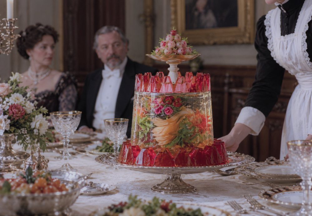 A server places a tiered, cylindrical gelatin mold filled with flowers and fruit on a formal dining table.