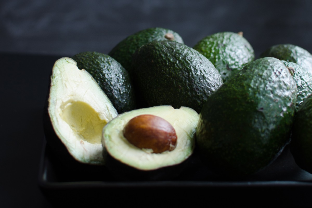 A pile of whole avocados next to one halved avocado with the pit still inside.