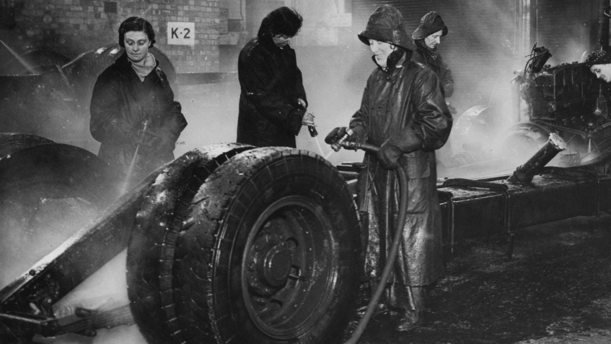 Four women in heavy coats and hats clean large vehicle wheels with hoses in a steamy, industrial setting. Brick walls and machinery are visible in the background, with a sign labeled “K-2” on the wall.
