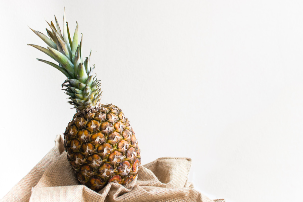 A whole pineapple resting on folded burlap fabric against a white background.