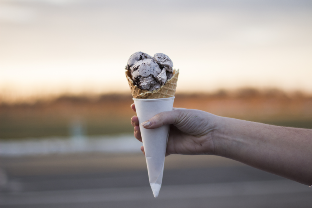 A hand holds a scoop of cookies and cream ice cream in a waffle cone wrapped in white paper.