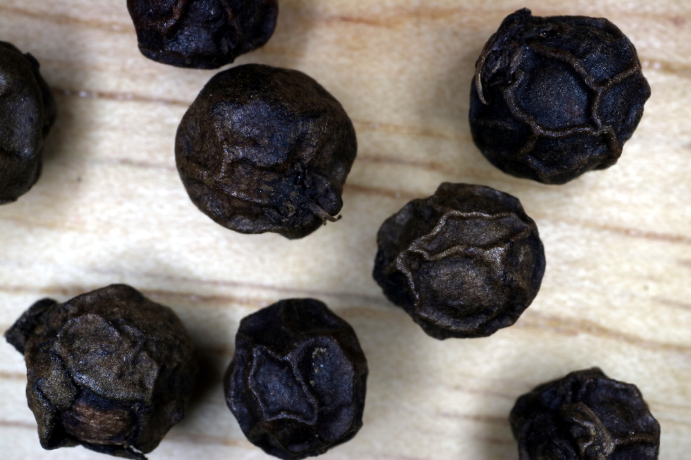 Seven whole black peppercorns scattered on a light-colored wooden surface.