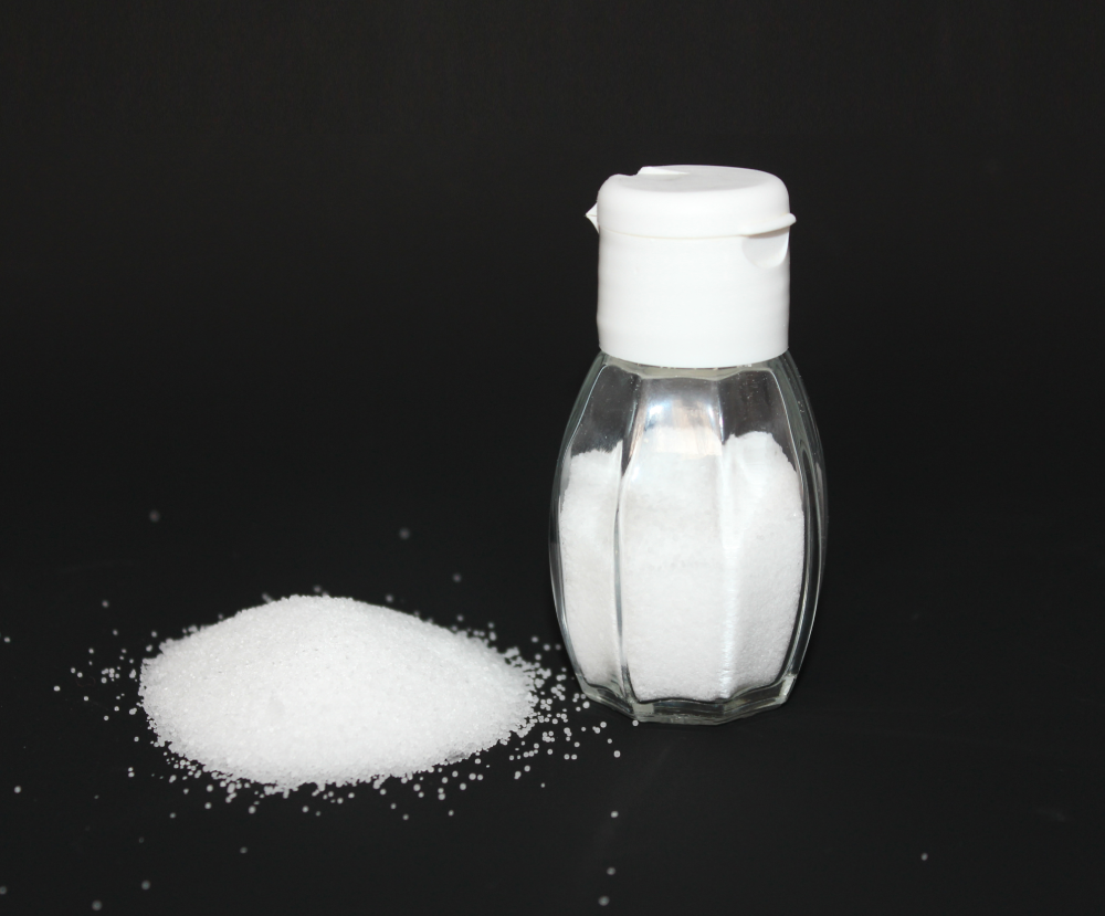 A glass salt shaker with a white cap sitting next to a small pile of salt on a black surface.