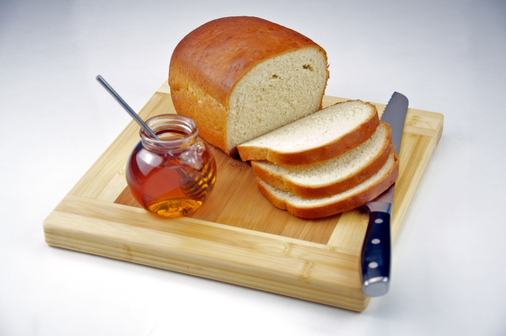 Loaf of bread with three slices, a jar of honey with a dipper, and a bread knife on a wooden cutting board.
