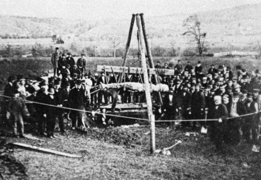 A large crowd of people standing in a field around a wooden tripod crane lifting a long, cylindrical object from the ground.