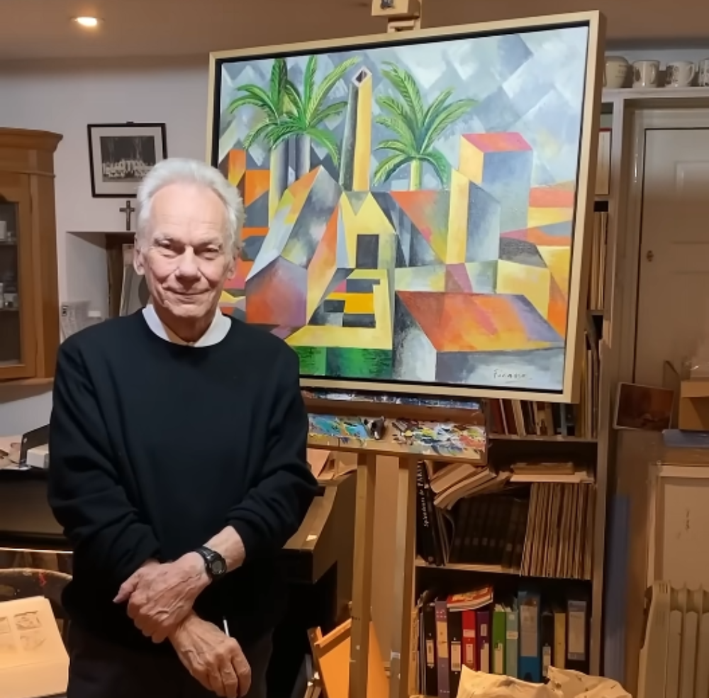 An older man standing in a studio with his hands clasped in front of an easel displaying a cubist-style painting of a colorful town with palm trees.