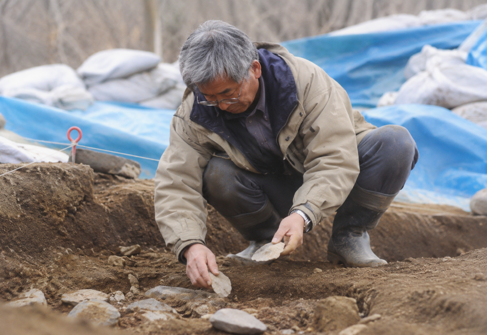 An archaeologist in a jacket and boots crouches at an excavation site, carefully examining stone artifacts in the dirt.