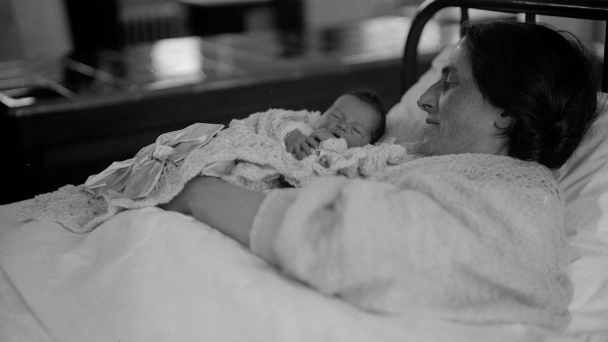 A woman lies in bed, holding a newborn baby wrapped in a blanket, both resting peacefully. The scene suggests a hospital or maternity ward setting.