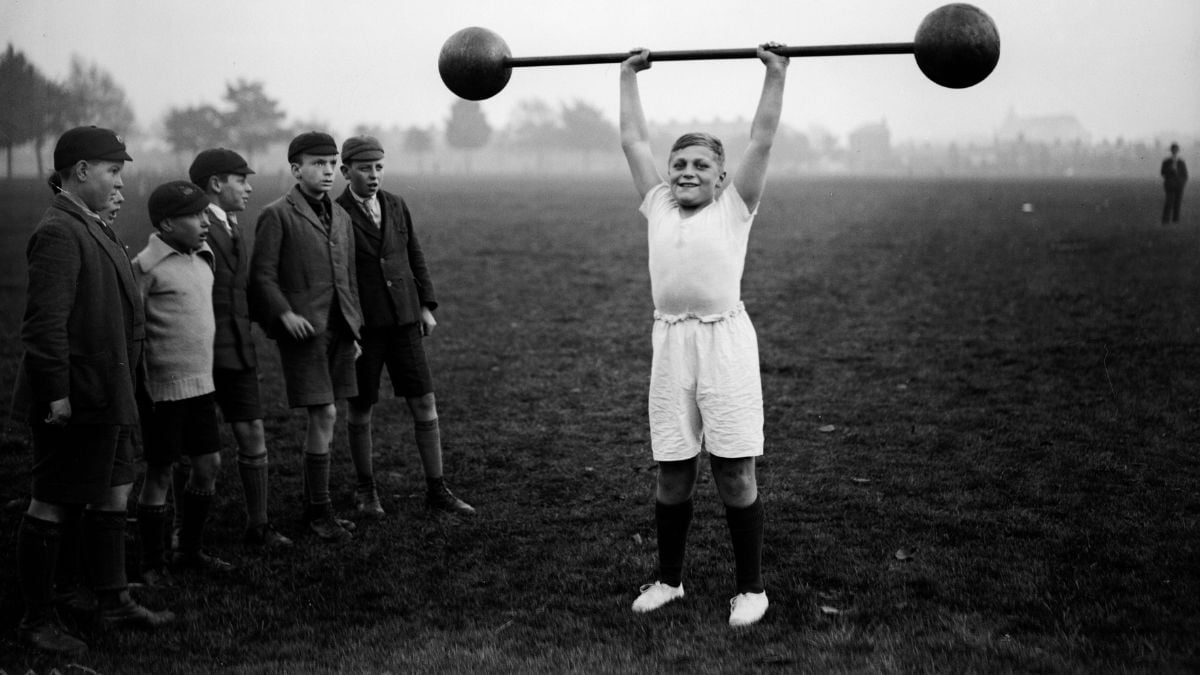 A boy in white shorts and shirt lifts a large, old-fashioned barbell overhead while a group of boys in hats and jackets watch on a grassy field. The scene appears vintage and outdoors.