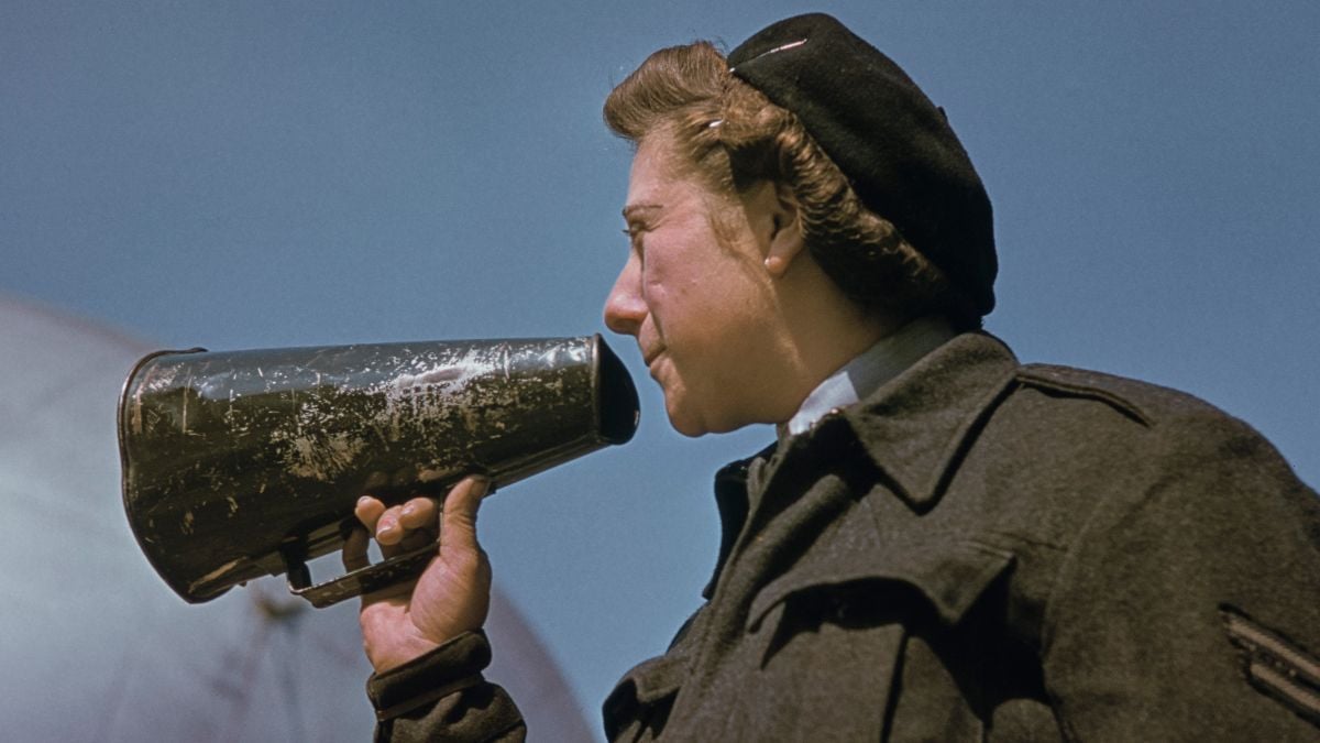 A woman in a dark coat and beret speaks into a weathered megaphone against a clear blue sky, viewed from a low angle.