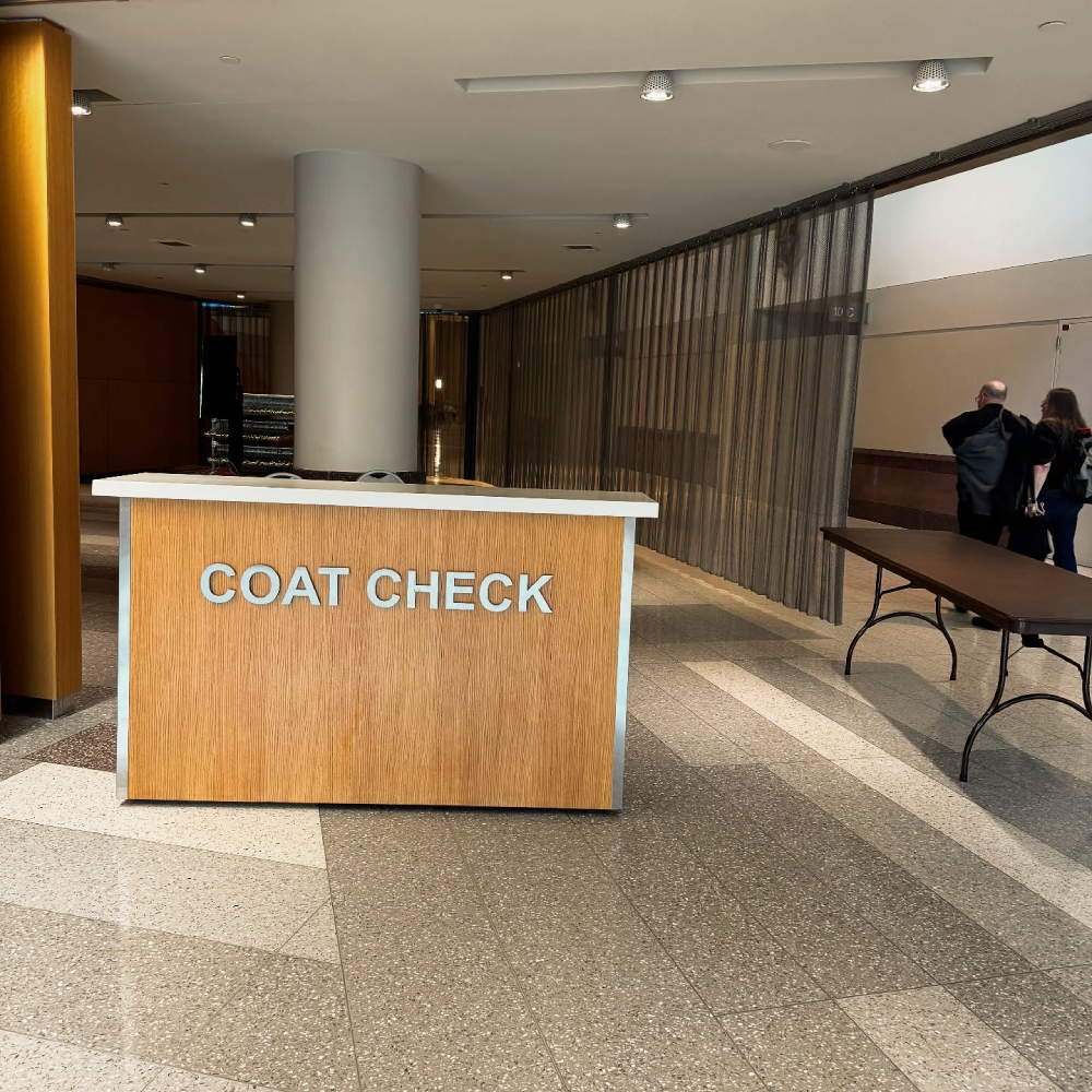 A wooden coat check desk with the words “COAT CHECK” in silver letters, situated in a spacious lobby with a metal curtain partition.