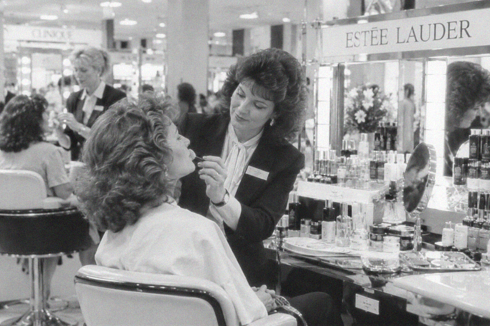 A makeup artist applies lipstick to a customer at an Estée Lauder beauty counter in a department store.