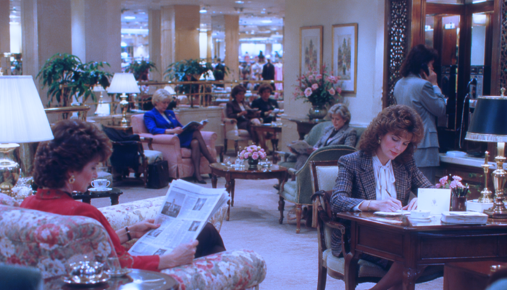 People sitting in a quiet, upscale department store lounge, with some reading newspapers and others working at desks.