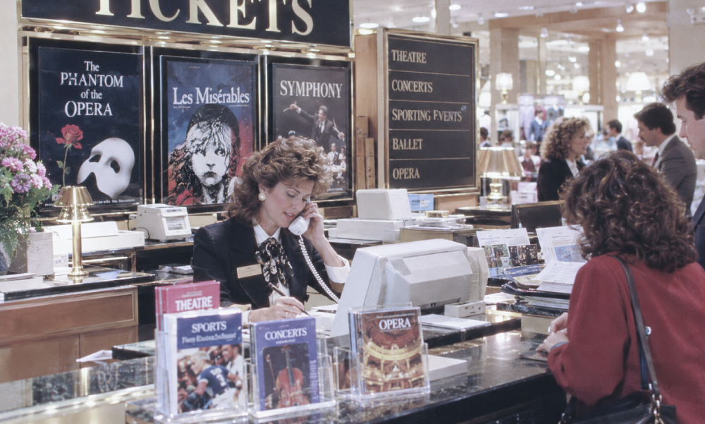 A ticket agent talks on the phone while assisting a customer at a service desk featuring posters for The Phantom of the Opera, Les Misérables, and symphony performances.